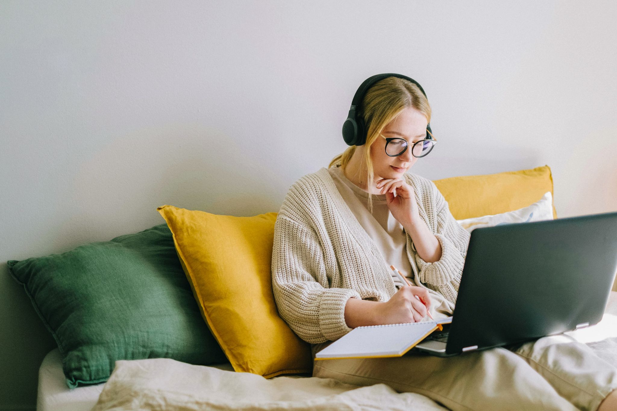 Viridian Central A woman in a cozy sweater and headphones sitting on a bed with a laptop, looking focused.